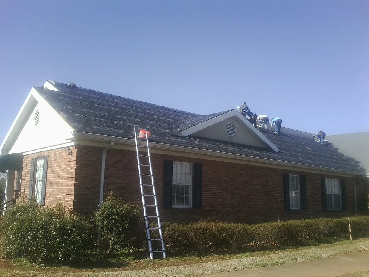 Crew tearing shingles off a brick ranch roof with a ladder set — Metal Roof Repair work by our crew in Mayer